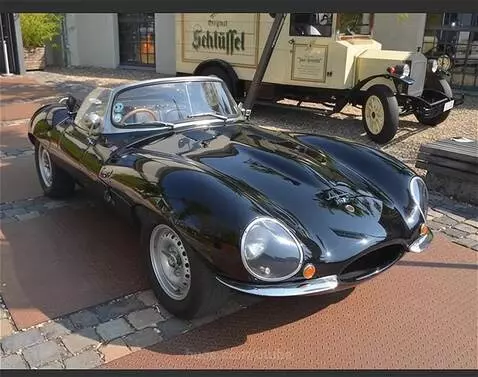 Polished black classic sports car parked outdoors, with a vintage cream delivery truck in the background.