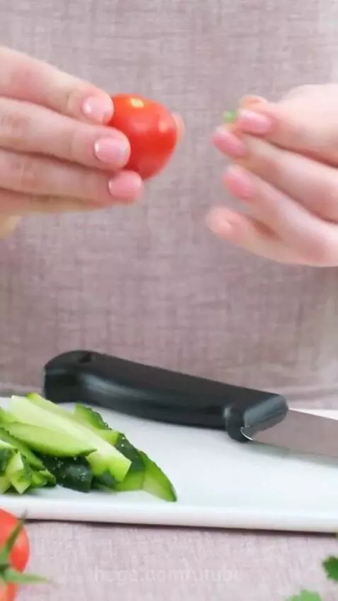 Close-up of hands slicing a red cherry tomato on a white cutting board with sliced cucumbers.