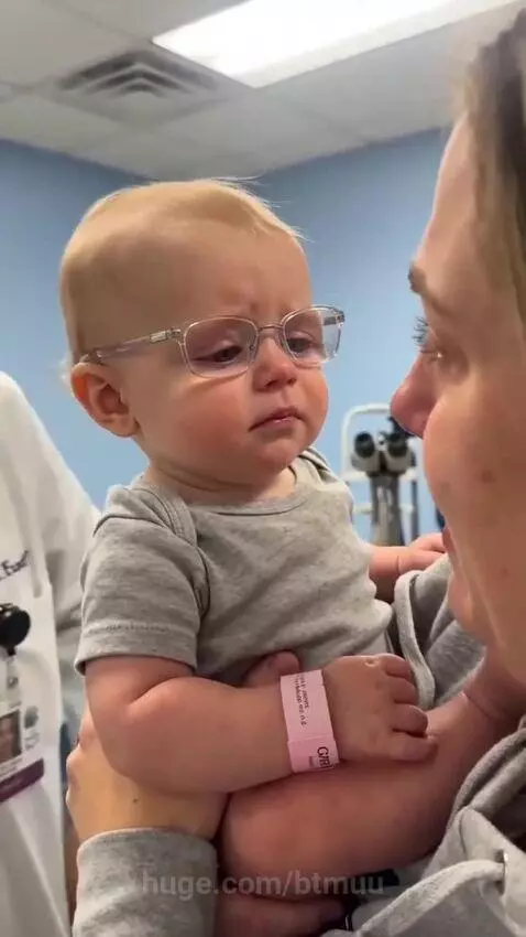 Baby with light hair wearing a grey onesie and a pink hospital band, looking confused as glasses are placed on their face, then smiling widely.