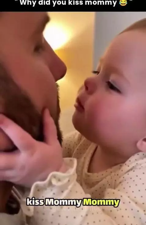 Baby with light hair and polka-dot top holds dad's cheek and asks why he kissed mommy.