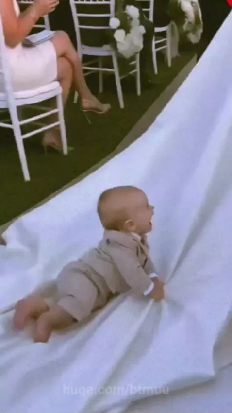 Baby in a suit crawling and playing with a bride's wedding dress train on an aisle.