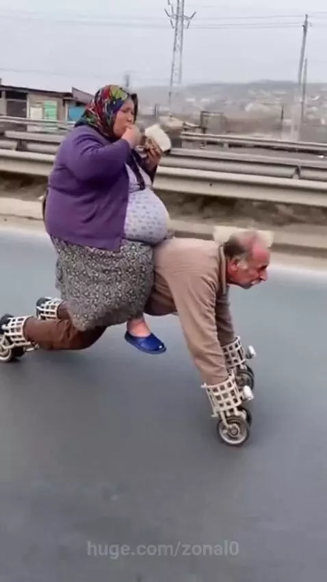 Elderly woman riding on an elderly man's back, both with wheels, falls onto a paved road.