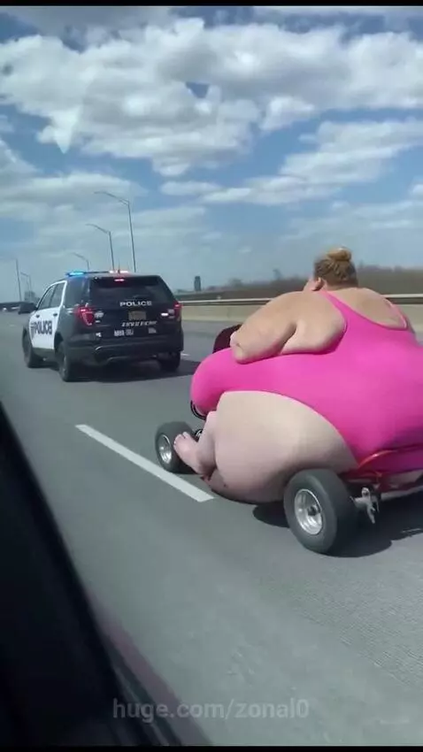 Woman in pink swimsuit riding a go-kart on a highway with a police SUV following.