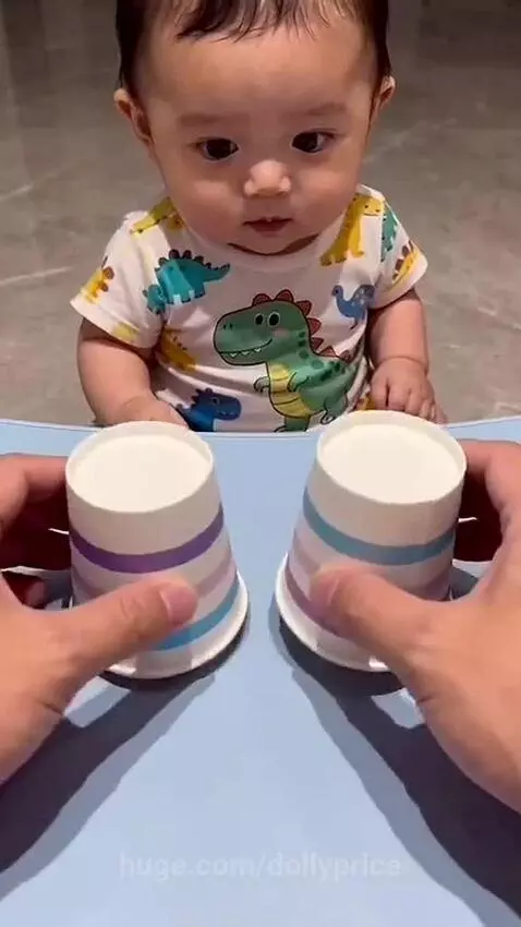 Baby in dinosaur onesie focused on a cup game, reacting with surprise to kibble reveal.