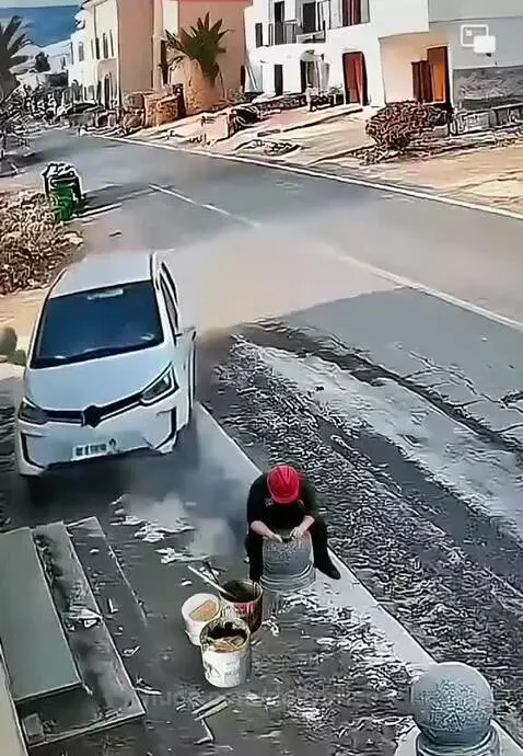 Person in red hat and dark clothes standing up from a bucket, splashing water next to a white car on a wet street.