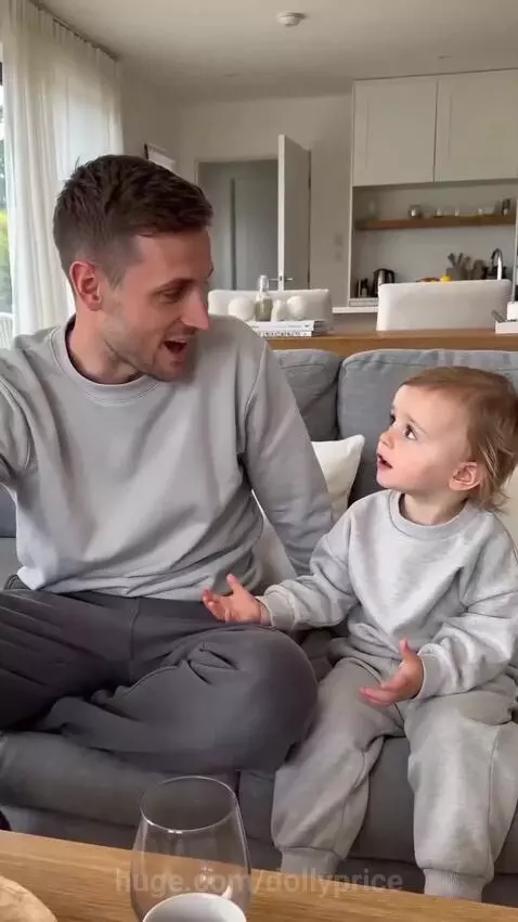 A young child in grey loungewear sits with her father on a couch, looking at him with a curious expression.