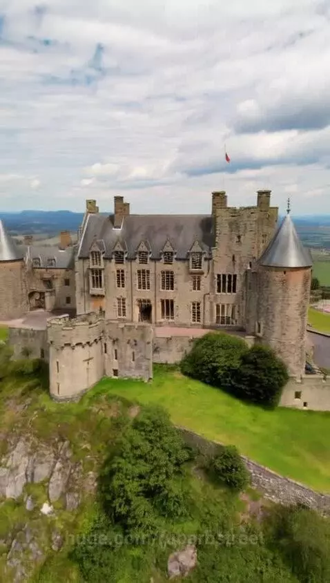 Aerial drone view of a large, historic castle on a grassy hill in the Scottish Highlands with rolling hills and a partly cloudy sky.