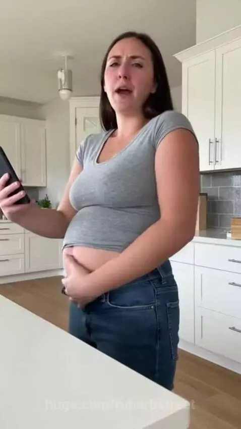 Woman holding her bloated stomach as her jeans button pops open in a kitchen.