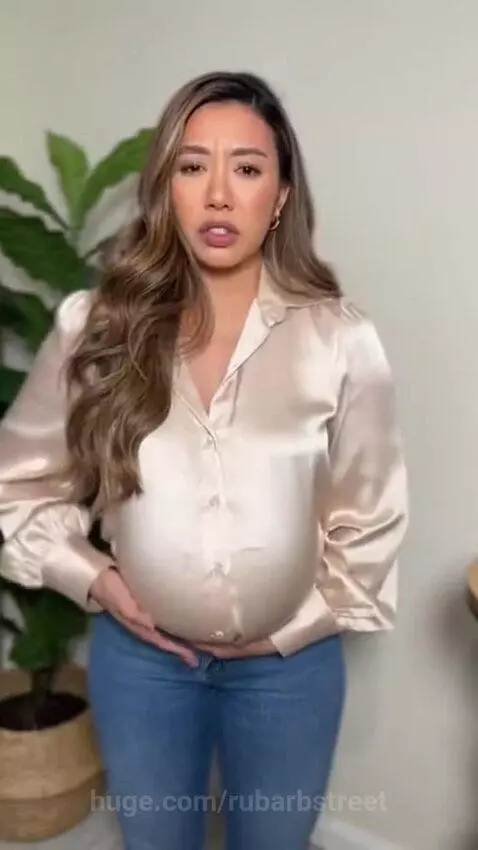 Woman with long brown hair in a satin blouse and jeans, looking surprised as her stomach visibly expands.