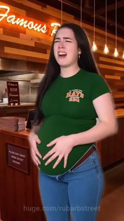Woman in green t-shirt and jeans holding distended stomach in apparent discomfort at a restaurant.