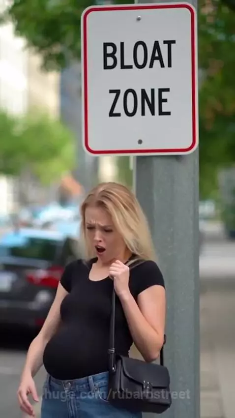 Woman in black t-shirt and jeans gasps as her stomach rapidly expands to a very large size next to a 'BLOAT ZONE' street sign.