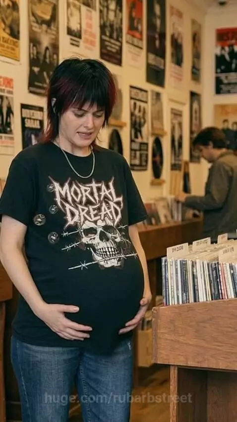 Record store owner with dark hair, wearing a 'MORTAL DREAD' t-shirt, holding her large, bloated belly with iridescent bubbles.