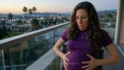 Woman with curly dark hair and glasses, wearing purple, holding distended abdomen in pain on a balcony overlooking a city at dusk.