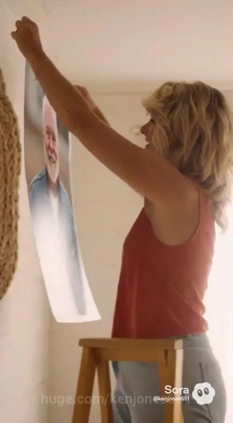 Woman with blonde hair in red top hanging a poster of a man with a white beard on a wall.