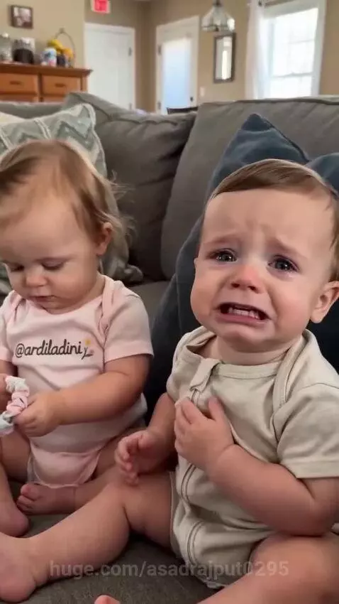 Baby girl in pink onesie holding scrunchie, baby boy in light onesie looking at her, with a crying voiceover.