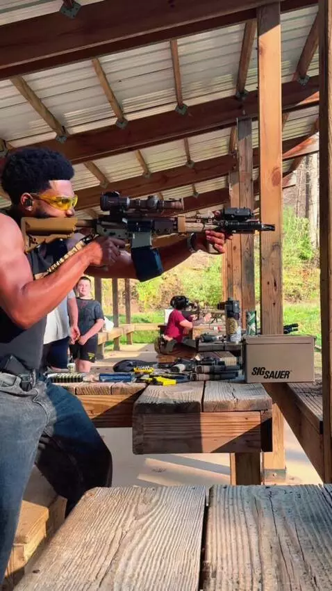 Man with afro and yellow safety glasses firing a rifle with a drum magazine at an outdoor shooting range.
