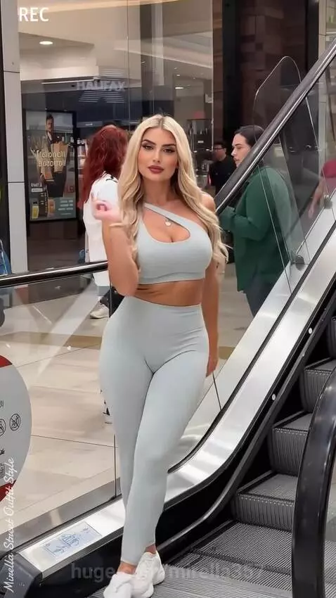 Blonde woman in blue sports bra and leggings dancing on a shopping mall escalator.