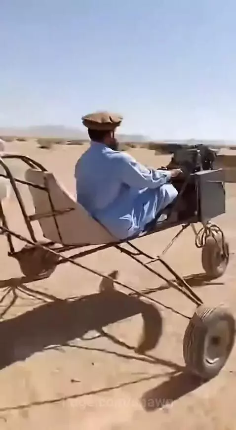 Man in traditional clothing flying a propeller-powered vehicle in a desert with onlookers cheering.