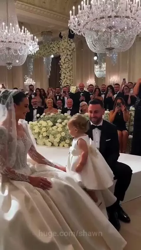 Flower girl sits on bride's lap at wedding, groom covers face playfully.