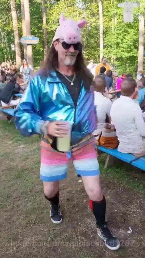 Man with long hair, pink pig hat, sunglasses, and iridescent jacket dancing outdoors with a drink.