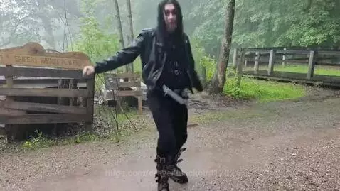 Person with long dark hair dancing energetically in the rain, wearing a black leather jacket and boots.