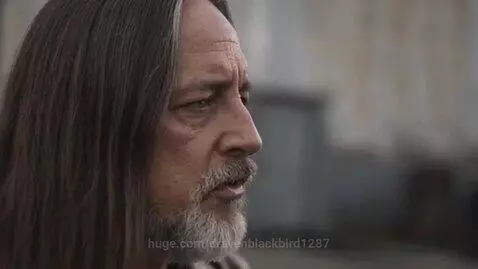 Man with long dark hair and beard in leather jacket singing in an outdoor industrial area with garage doors.