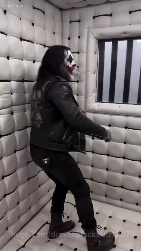 Man with clown makeup and skull jacket in a padded room, expressing distress.
