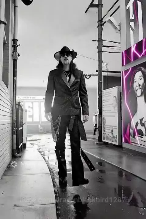 Man with long hair, beard, sunglasses, and hat in a double-breasted suit walks towards camera in a smoky, neon-lit urban environment.