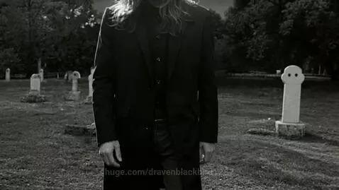 Man with serious expression walks slowly towards camera in a black and white graveyard with gravestones.