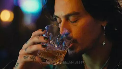 Man with beard and leather jacket sitting at a crowded bar, drinking from a glass.