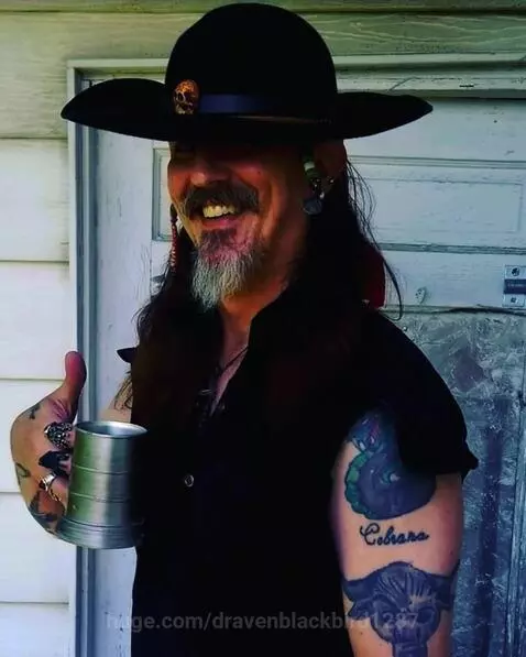 Man with pirate hat, beard, tattoos, and a mug, smiling.