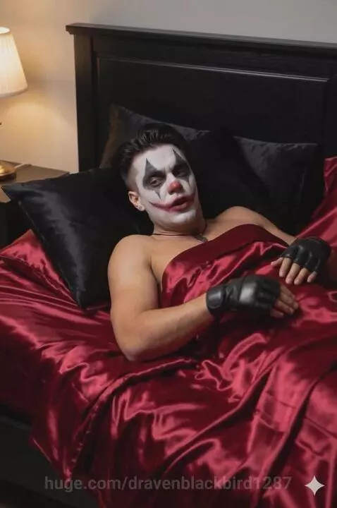 Clown with dramatic makeup and fingerless gloves waking up in a bed with red satin sheets.