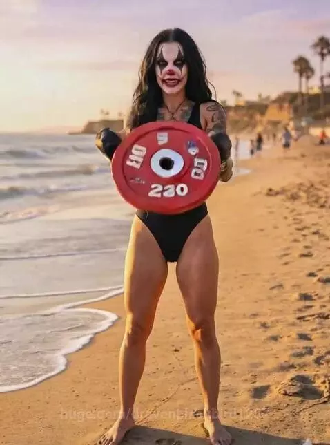 Woman with clown makeup and boxing gloves performing a front raise with a weight plate on a sandy beach.