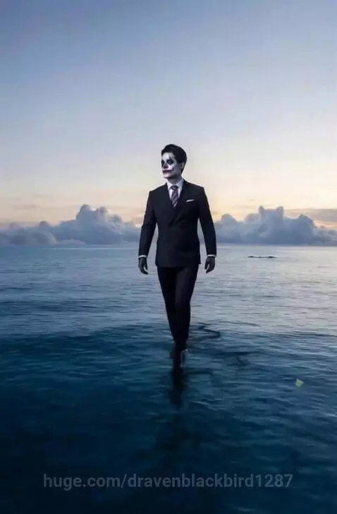 Man with skull face paint walking on the surface of a calm blue ocean with a colorful sunset sky.