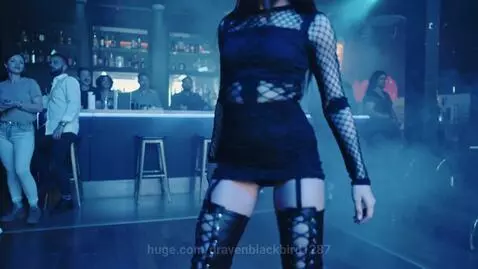 Woman in black dress and thigh-high boots dancing suggestively in a dimly lit club with blue and purple lighting.