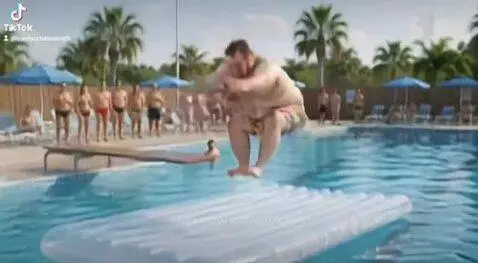 Overweight man in swim trunks diving from a board, landing on an inflatable mattress with a large splash.