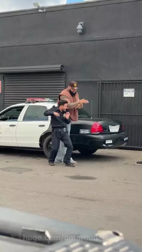 A small police officer attempting to handcuff a much larger man, with a police car in the background.