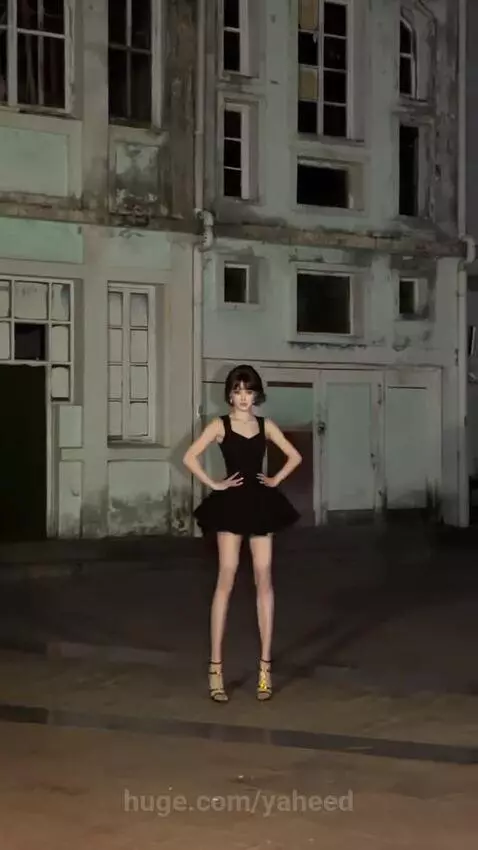 Woman with short dark hair in a black mini-dress and gold heels dancing energetically outdoors at night.