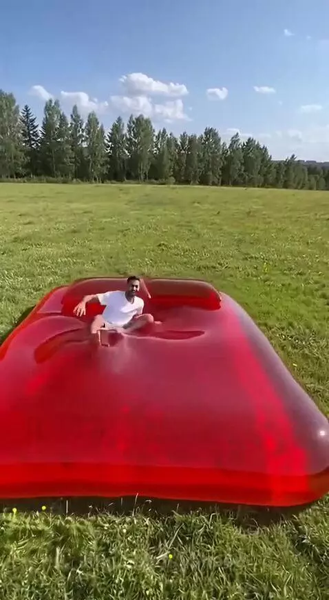 Man jumping on a large, red, transparent inflatable mattress in a grassy field.