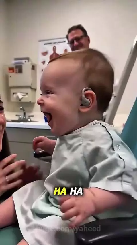 Close-up of a baby smiling and laughing while wearing hearing aids, reacting to a parent's voice.