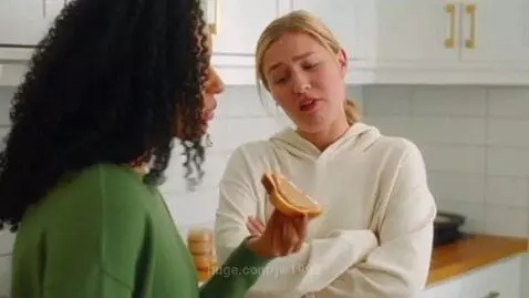 Woman with peanut butter toast playfully withholding it from a friend in a bright kitchen.