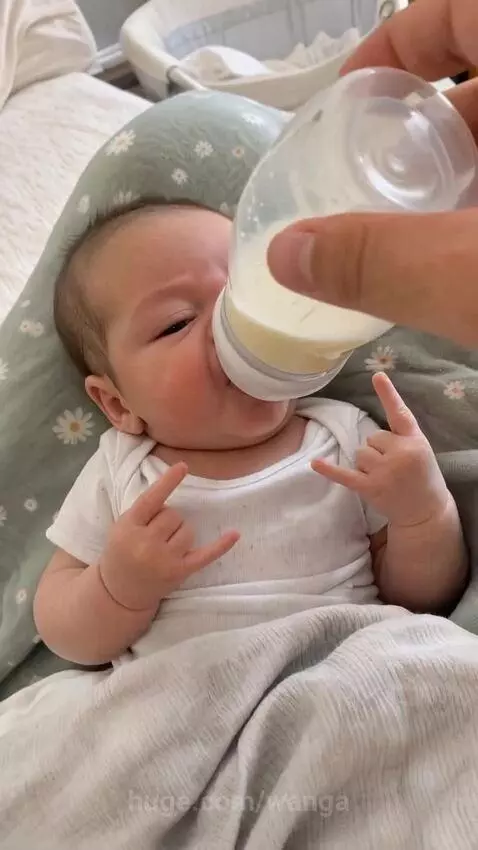 Baby in a onesie lying down, bottle-feeding and making the rock and roll hand sign.