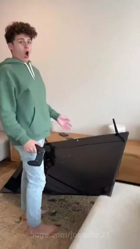 Barefoot man in green hoodie looking shocked at a shattered flat-screen TV on the floor, holding a game controller.
