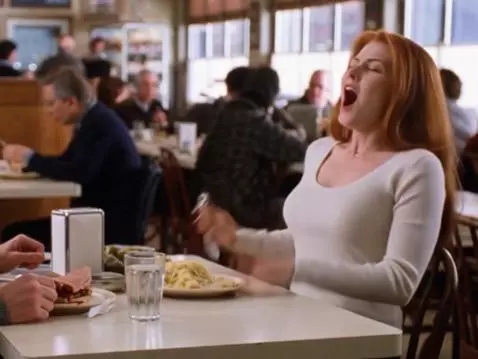 Woman with long red hair faking an orgasm at a diner table while a man looks on surprised.