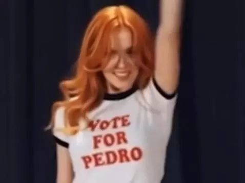 Woman with long red hair dancing energetically in a 'Vote for Pedro' shirt and black mini-skirt on a wooden stage.