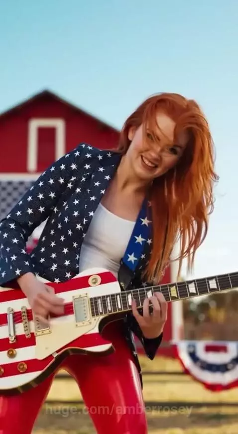 Woman with red hair in a star-spangled blazer and red pants plays an American flag electric guitar, smiling.