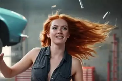 Woman with red hair in grey top and denim shorts dancing in a garage with a car on a hoist and floating tools.