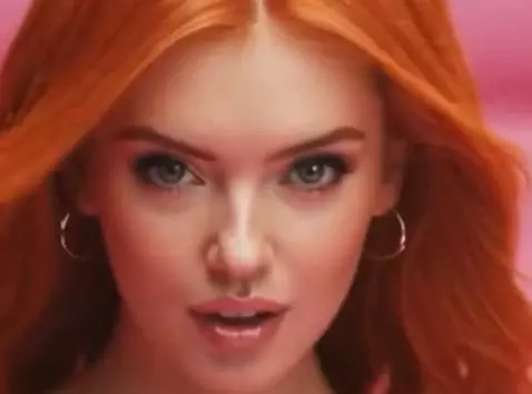 Woman with long red hair in a pink satin dress and heels on a pink set, posing confidently.