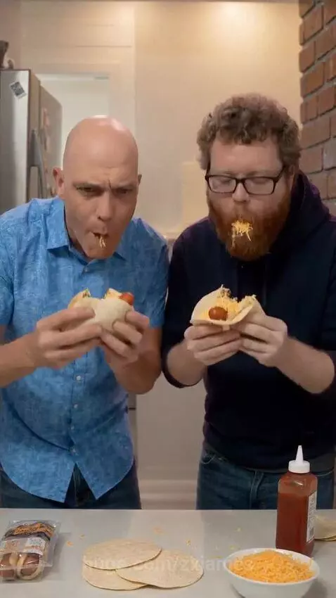 Two men laughing while making and eating messy hotdog tacos with a large mustard spill.