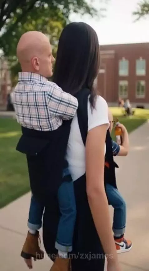Woman walking outdoors carrying two bald men in baby carriers, one on front, one on back.